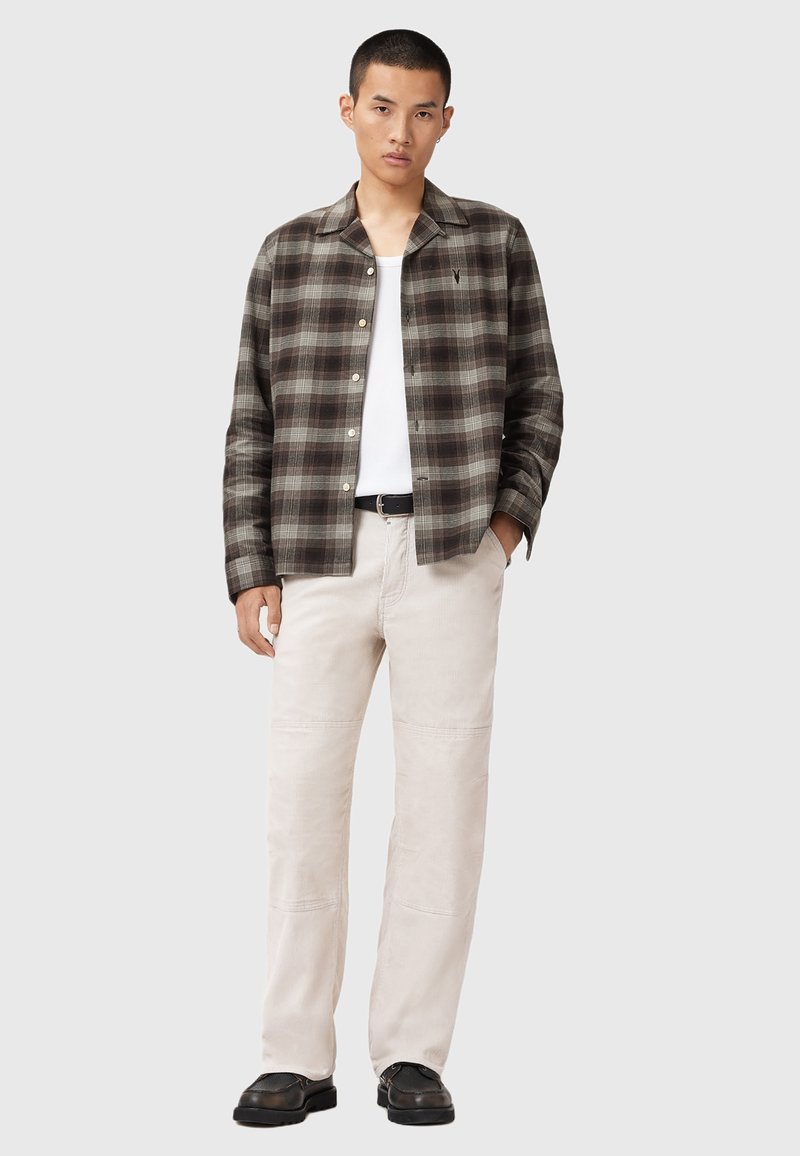 Young man wearing brown and gray plaid button-up shirt, white tank top, beige pants, black belt, and black shoes standing with one hand in pocket.