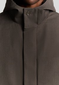 Donkerbruine jas met hoge kraag, gladde textuur en een enkele metalen knoopdetail. Minimalistisch ontwerp met een getailleerde pasvorm.