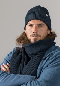 Dark blue ribbed knit beanie and matching scarf worn over a blue fleece jacket. The scarf features a textured pattern. Model stands arms crossed.