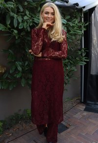 Burgundna čipkasta obleka z rožnim vzorcem, dolgimi rokavi, rahlo prosojnim materialom in dolgim robom. Nošena čez oprijete hlače.