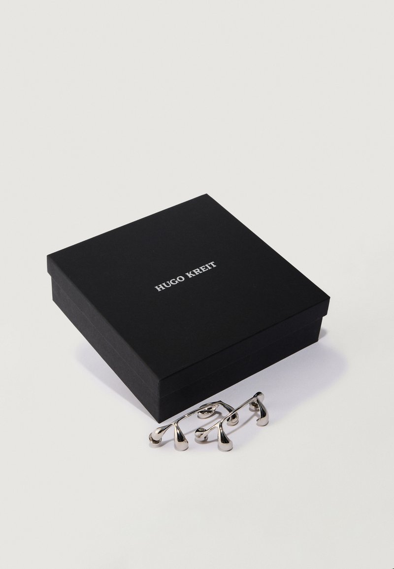 Schwarze Hugo Kreit Box neben einem Paar silberner Manschettenknöpfe mit abgerundeten Enden auf einer weißen Oberfläche.
