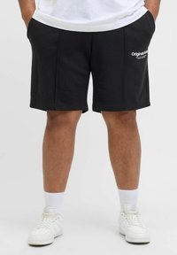 Shorts en coton noirs avec poches latérales, jusqu'aux genoux, ornés d'un logo blanc sur le côté gauche et d'une texture lisse. Portés avec des baskets blanches.