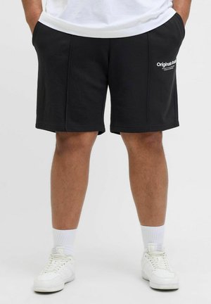 Svarta bomullsshorts med sidofickor, knälånga, med en vit logotyp på vänster sida och en slät yta. Bärs med vita sneakers.