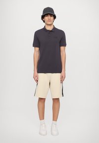 Tmavošedé polo tričko s krátkym golierom, béžové šortky s čiernymi bočnými pruhmi a biele tenisky. Neformálny outfit s jednoduchými dizajnovými prvkami.