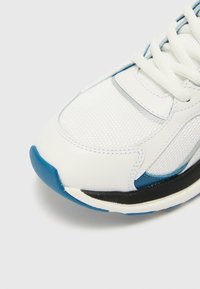 White sneaker featuring mesh and synthetic materials, with a blue toe cap, grey accents, and black outsole. Laces are white and flat.