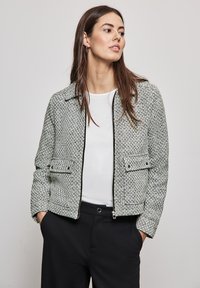 Textured jasje in een zwart-wit houndstooth patroon, met een rits aan de voorkant, twee voorkeurszakken en een kraag. G gedragen over een witte top.
