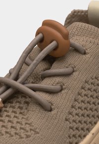 Κοντινό πλάνο σε μπεζ πλεκτό αθλητικό παπούτσι με ελαστικά κορδόνια ασφαλισμένα με καφέ κούμπωμα στην υφασμάτινη επάνω επιφάνεια του παπουτσιού.