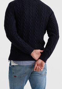 Mörkblå kabelstickad tröja med ribbad nederkant och ärmslut, bärs över en ljusblå rutigt skjorta, tillsammans med ljusa denimjeans.