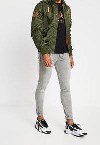 Hombre con una chaqueta bomber verde oliva, camiseta gráfica negra, vaqueros ajustados gris claro y zapatillas blanco y negro, de pie contra un fondo blanco.