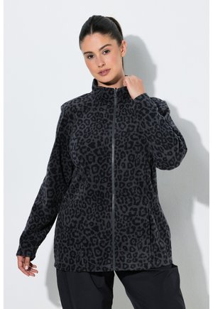 Kvinde iført en sort og mørkegrå leopardprint fleecejakke med lynlås og lange ærmer, stående foran en ensfarvet hvid baggrund.