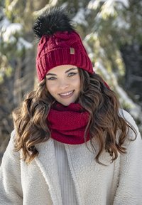 Gorro de punto rojo con diseño acanalado y pompón de pelaje sintético negro, acompañado de una bufanda infinita roja texturizada a juego. Abrigo blanco forrado de forro polar.