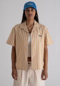 Striped short-sleeve shirt in cream and orange with a collared neckline. Wearing a blue cap with embroidered logo. Light-colored pants.