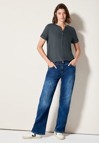 Camicia a maniche corte grigio scuro, con colletto e vestibilità rilassata, abbinata a jeans a gamba larga in denim chiaro e sneakers bianche con dettagli neri.