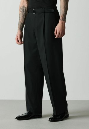 Man wearing black wide-leg pants with a belt and shiny black dress shoes, standing against a plain background.