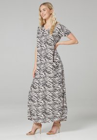 Maxi-Kleid mit V-Ausschnitt und kurzen Ärmeln in Beige, das ein schwarzes wellenförmiges Muster aufweist. Leichter Stoff, bodenlang, kombiniert mit goldenen Sandalen.