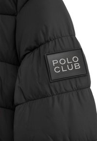 Chaqueta acolchada negra con una superficie texturizada, con una etiqueta rectangular negra grabada con "POLO CLUB" en letras grises en la manga.