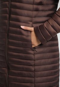 Braune Steppjacke mit horizontaler Unterteilung, ausgestattet mit einem Reißverschluss und seitlichen Taschen. Glattes, glänzendes Material mit gepolsterter Textur.