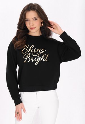 Ung kvinde med langt brunt hår iført sort sweatshirt med guldteksten "Shine Bright" og hvide bukser, poserer mod en hvid baggrund.