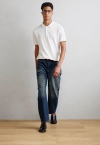 Polo bianco, jeans blu scuro con sfumature, e scarpe nere. Il modello indossa occhiali e si trova di fronte a uno sfondo grigio chiaro.