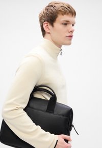 Black leather bag with textured surface, rectangular shape, dual top handles, and zip closure, held against light beige top.