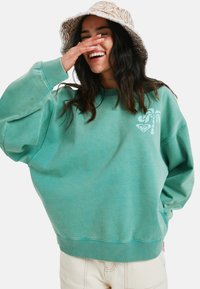 Tealowy, oversize sweter z ribbami przy mankietach, graficzny wzór palm na klatce piersiowej oraz miękka, strukturalna tkanina. Uzupełniony wzorzystą czapką-bucket.