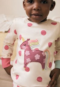 Haut blanc à manches longues avec des pois roses et une application de licorne rose dotée d'une crinière multicolore et de détails floraux, tissu doux.