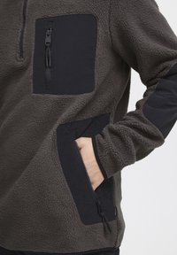 Fleece-pullover i mørkegrå med svarte detaljer; har en halv-zip foran, to glidelåslommer og teksturert stoff.