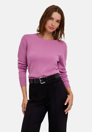 Femme portant un pull rose à manches longues rentré dans un pantalon noir taille haute avec une ceinture noire, posant devant un fond clair uni.