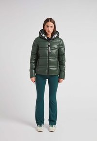 Chaqueta acolchada verde con un acabado brillante, cierre de cremallera y una capucha negra con un logo blanco. Combinada con pantalones azul claro y zapatillas blancas.