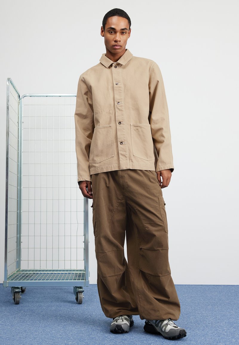 Chaqueta de lienzo beige con cuello, cierre de botones y dos bolsillos frontales, combinada con pantalones cargo marrones de corte holgado y zapatillas grises.