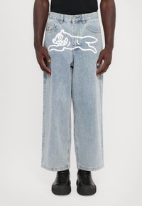 Jeans de perna larga em azul claro feitos de denim, com uma impressão de gato em estilo cartoon branco na coxa. Sapatos pretos de atacador exibidos abaixo.