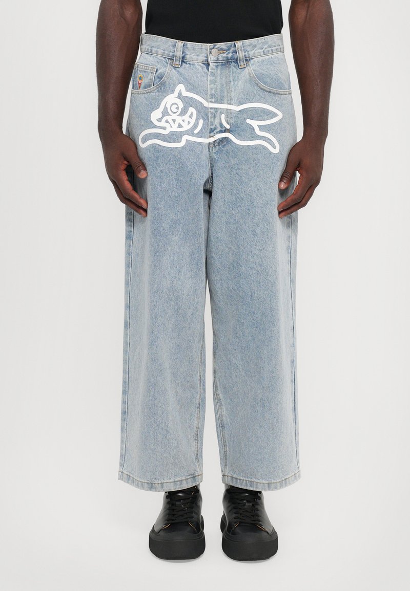 Jeans de perna larga em azul claro feitos de denim, com uma impressão de gato em estilo cartoon branco na coxa. Sapatos pretos de atacador exibidos abaixo.
