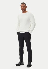 Uomo in piedi con le mani nelle tasche, che indossa un maglione bianco, pantaloni neri e scarpe eleganti nere su uno sfondo bianco.