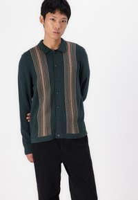 Cardigan verde scuro con strisce verticali color beige e motivo a trecce. Caratterizzato da un colletto e chiusura con bottoni. Tessuto morbido.