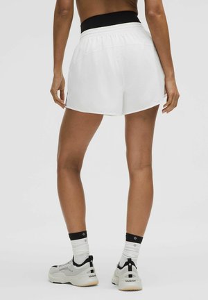 Persona de pie vistiendo pantalones cortos deportivos blancos, camiseta deportiva negra, calcetines blancos y zapatillas blancas con detalles en negro.