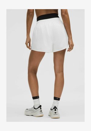 Persona de pie vistiendo pantalones cortos deportivos blancos, camiseta deportiva negra, calcetines blancos y zapatillas blancas con detalles en negro.