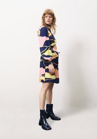 Mehrfarbiges Kleid mit abstrakten Mustern in Marineblau, Pink, Gelb und Grün; lange Ärmel, hoher Ausschnitt und lockere Passform, kombiniert mit schwarzen Knöchelstiefeln.