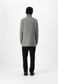 Cappotto in misto lana grigio con colletto alto, spacco posteriore e design sartoriale, abbinato a pantaloni e scarpe nere.