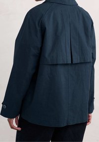 Osoba w ciemnoniebieskiej kurtce z długim rękawem i przypiętymi mankietami, widoczna od tyłu na jednolitym tle.