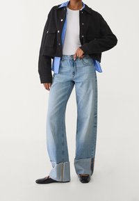 Osoba v světle modrých širokých džínách s ohrnutými manžetami, černé krátké bundě přes modrou košili, bílé tričko a černé boty.