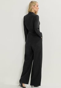 Combinaison noire avec un design ajusté, manches longues, jambes larges et poches. Texture douce, cintrée à la taille, avec un minimum de détails.