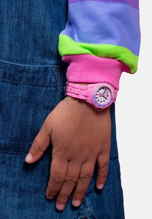 Orologio in silicone rosa al polso, con un quadrante bianco e indicatori colorati. Primo piano sulla mano su uno sfondo in denim blu.