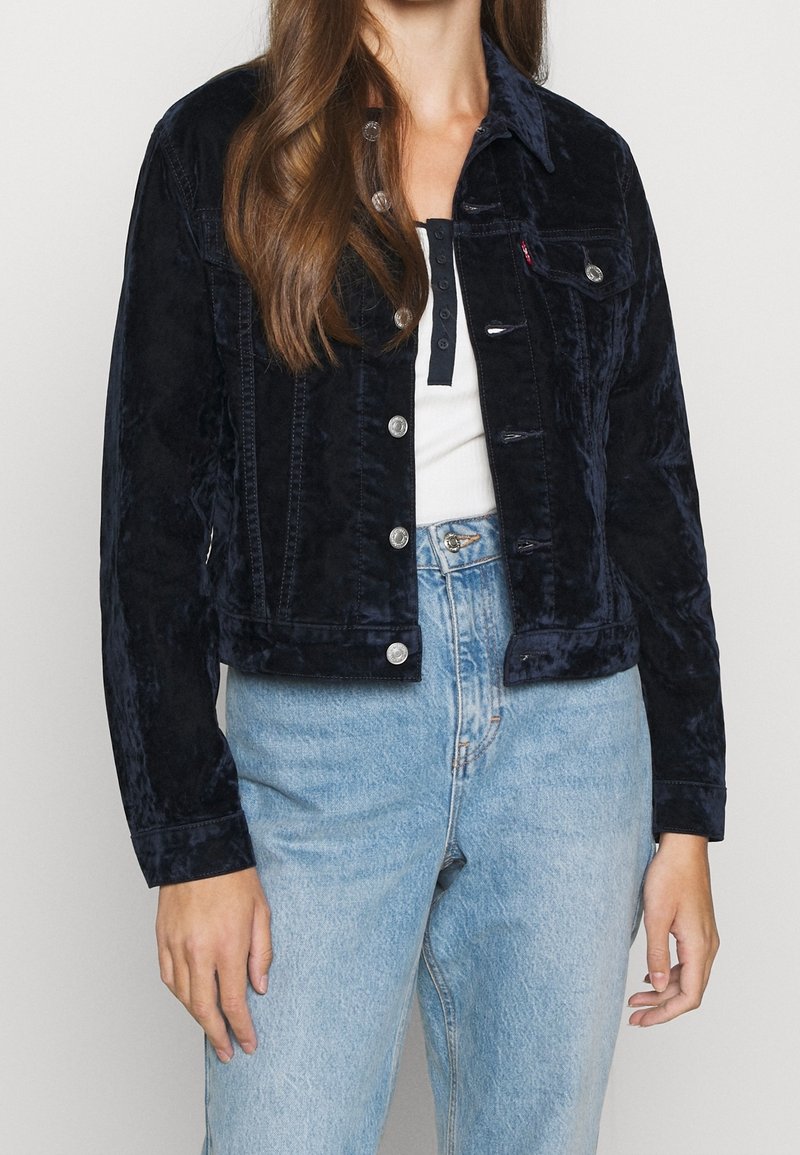 Femme portant une veste en velours bleu foncé à boutons par-dessus un haut blanc et un jean taille haute bleu clair, debout devant un fond uni.