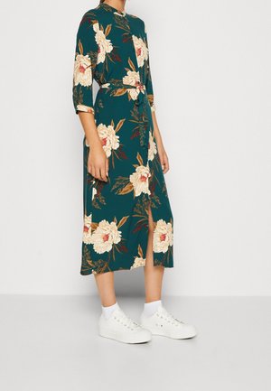 Femme portant une robe midi verte à fleurs avec une fente à l'avant, des manches trois-quarts, une taille nouée et des baskets blanches avec des chaussettes blanches.