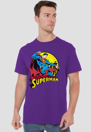 Young man wearing a purple t-shirt featuring Superman flying with cityscape and yellow sunset background.