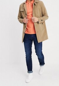 Béžový trenčkot přes oranžové polo tričko, doplněný modrými džíny a bílými teniskami. Trenčkot má knoflíky a klasický límec.