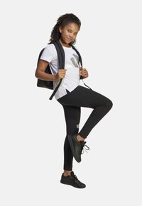 Mochila negra, camiseta blanca con gráfico metálico, leggings negros y zapatillas negras. Cabello rizado recogido en una coleta.