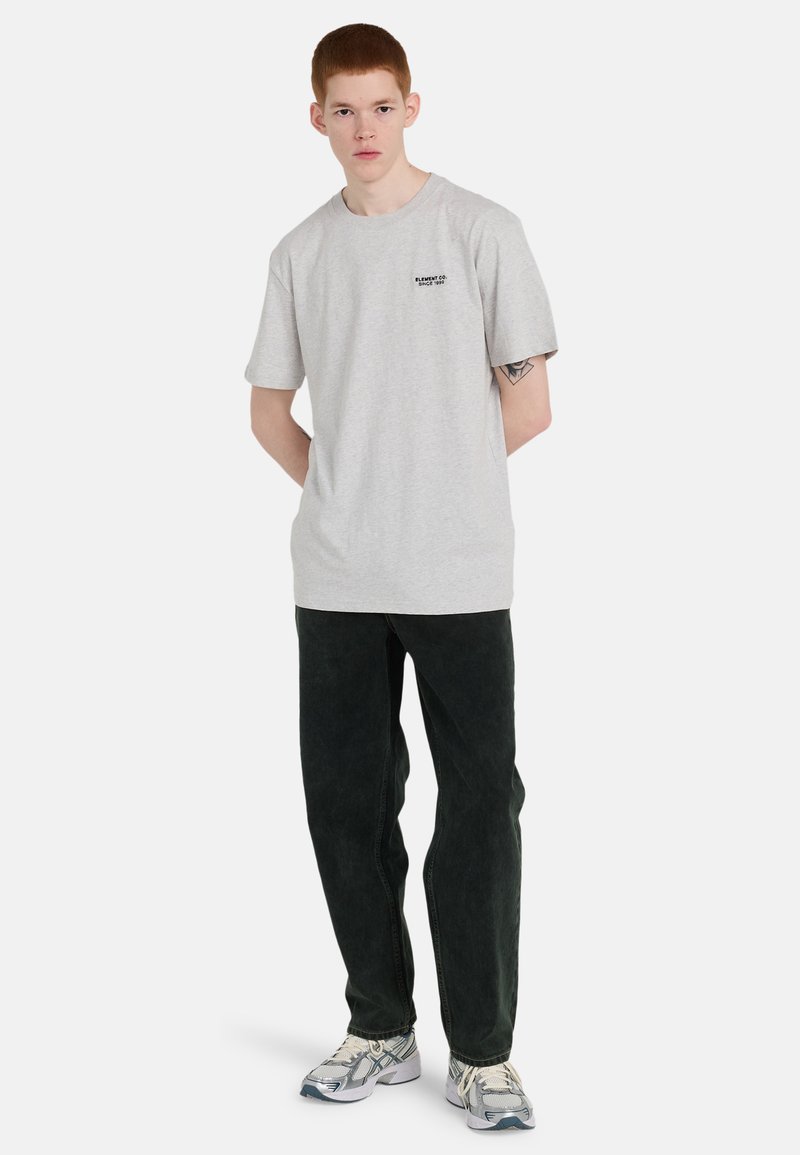 Jeune homme debout, mains dans le dos, portant un T-shirt gris clair, un jean noir, des baskets blanches et grises, avec un tatouage visible sur le bras.