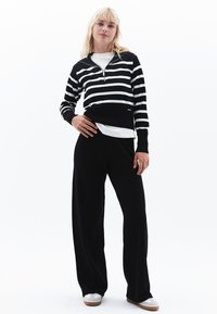 Maglione zip-up a righe bianche e nere abbinato a pantaloni larghi neri. Il maglione presenta polsini a coste e una maglietta bianca a contrasto sotto.