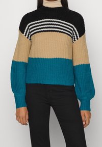 Frau trägt einen gestrickten Pullover mit schwarz-weißen Streifen sowie beige und türkisfarbenen Farbkombinationen, kombiniert mit schwarzen Hosen.